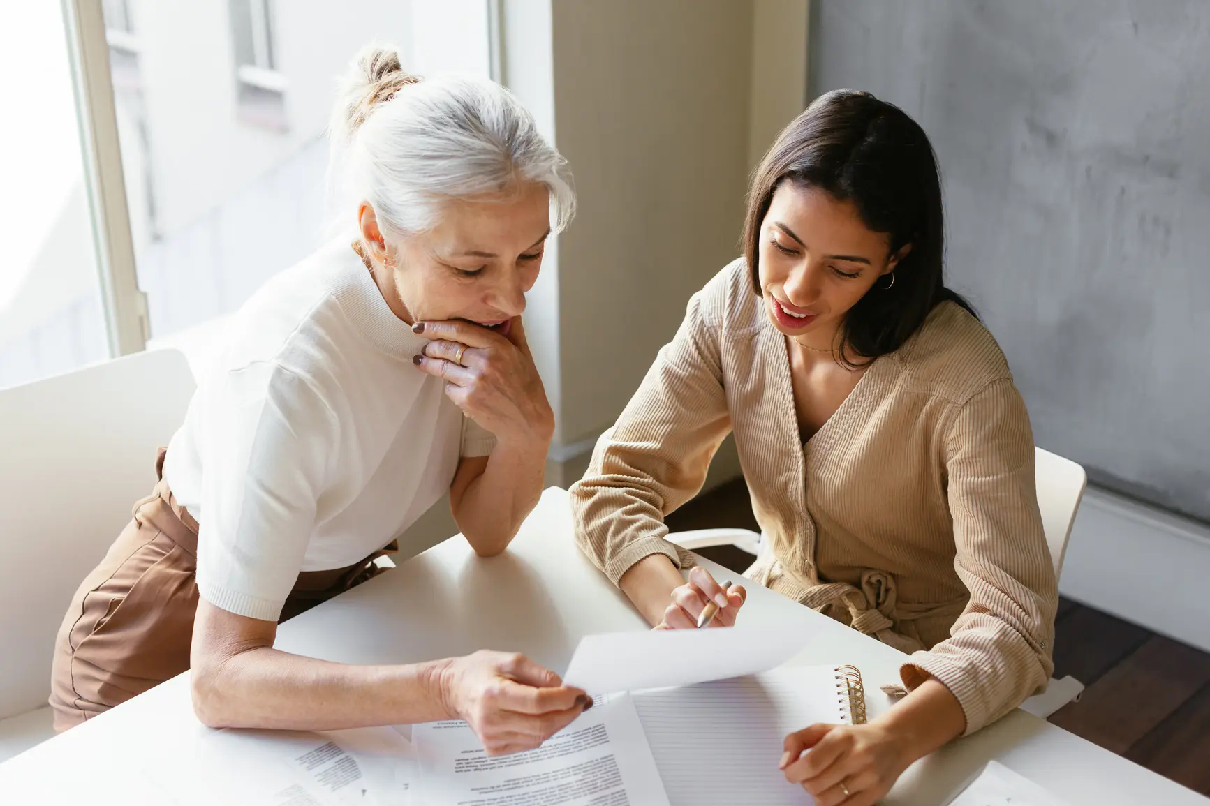 Two women are sitting at a table and looking at documents together.