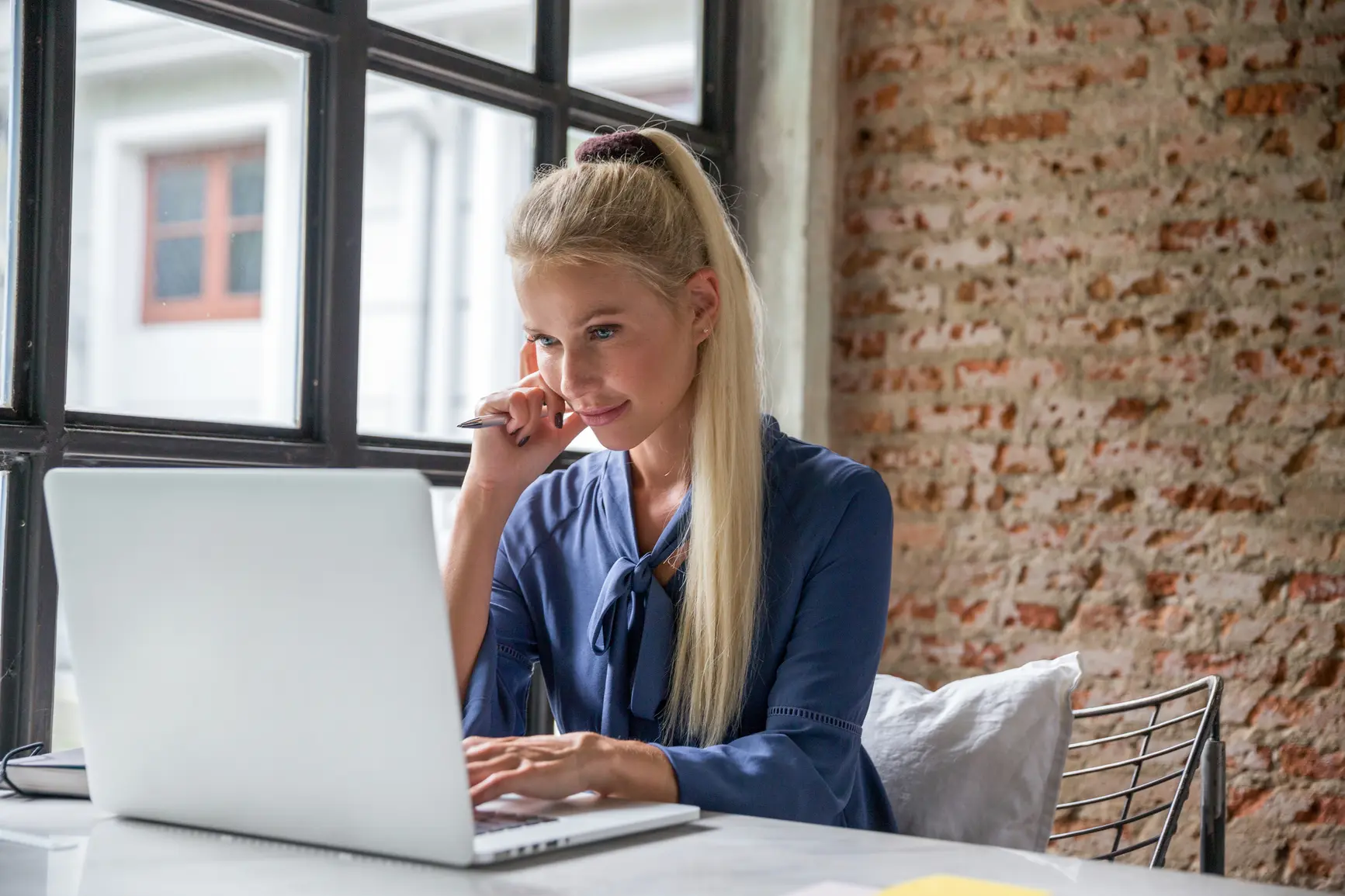 Eine junge Frau sitzt mit einer Hand am Laptop, einer Hand am Gesicht vor ihrem Laptop. Im Hintergrund ist eine Mauerwand zu sehen.