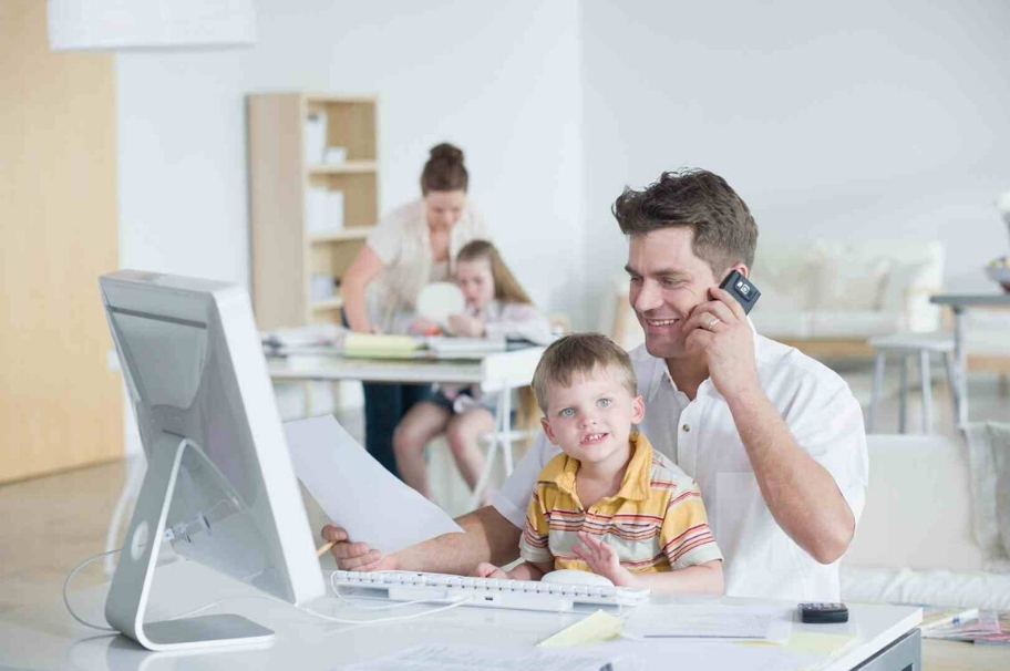 A man sits with his son on his lap at the laptop and talks on the phone. In the background you can see a woman working at a desk with a girl