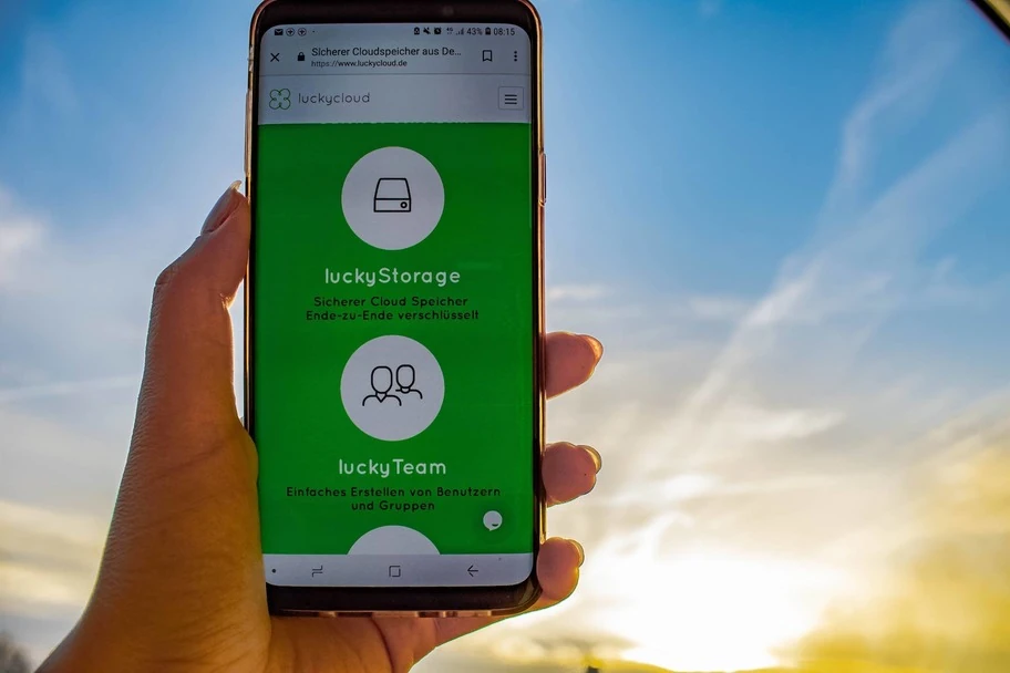 Ein Smartphone wird in die Hoehe gehalten - auf dem Bildschirm ist der luckyStorage zu sehen, im Hintergrund ein Sonnenaufgang.
