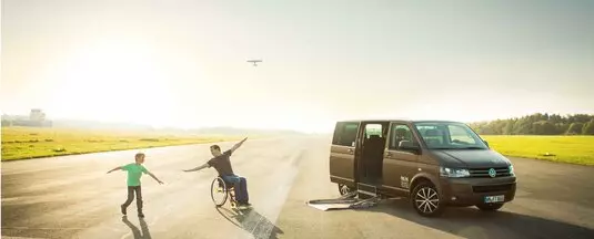 A man in a wheelchair romps with his son in front of a vehicle that has been modified to accommodate the disabled.